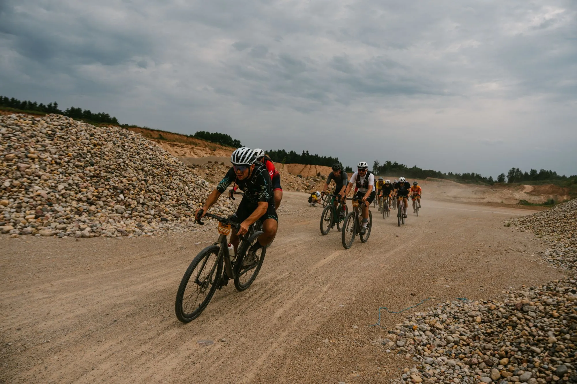 Gravel race through a sand quarry: a rider wearing number 507 leads a dusty line of cyclists past heaps of stones under a heavy grey sky.
